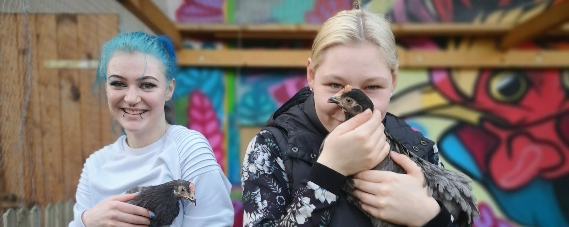 Zwei jugendliche Mädchen halten lächelnd je ein schwarzes Huhn auf dem Arm