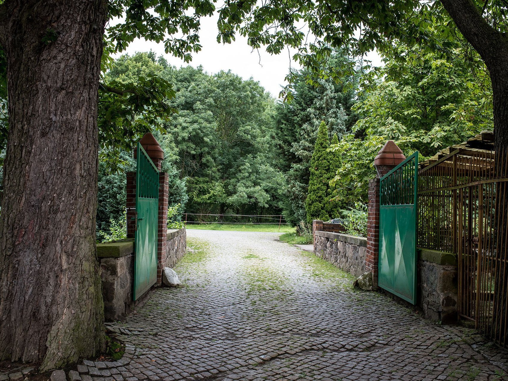 Unter Bäumen führt ein Kiesweg durch ein grünes Tor. Im Hintergrund Bäume